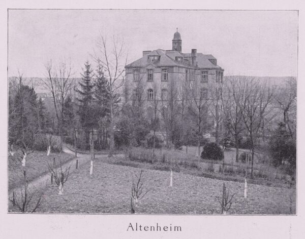 Historisches Foto des Altenheimgebäudes unweit des Bahnhofs. Vor dem mehrstöckicken Gebäude erstreckt sich ein Garten mit jungen Obstbäumen. Um das Gebäude herum sind weitere, gröere Bäume. Im Hintergrund liegt die Stadt Idstein.