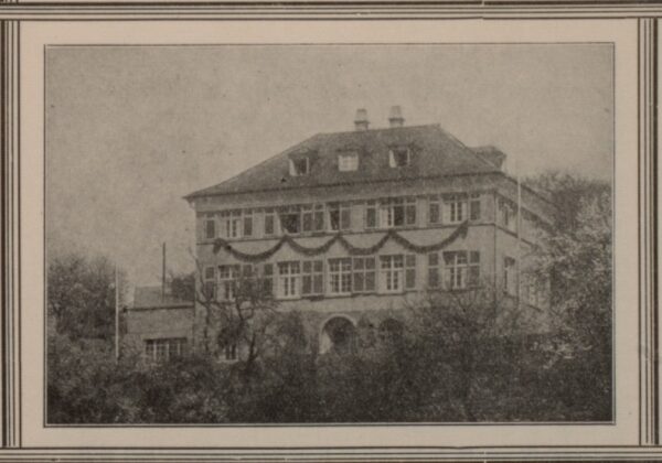 Historisches Foto des Krankenhausgebäudes.Dreistöckiges Gebäude, im Vordergund sind Bäume. Im oberen Stockwerk schmücken Girlanden die Fassade.